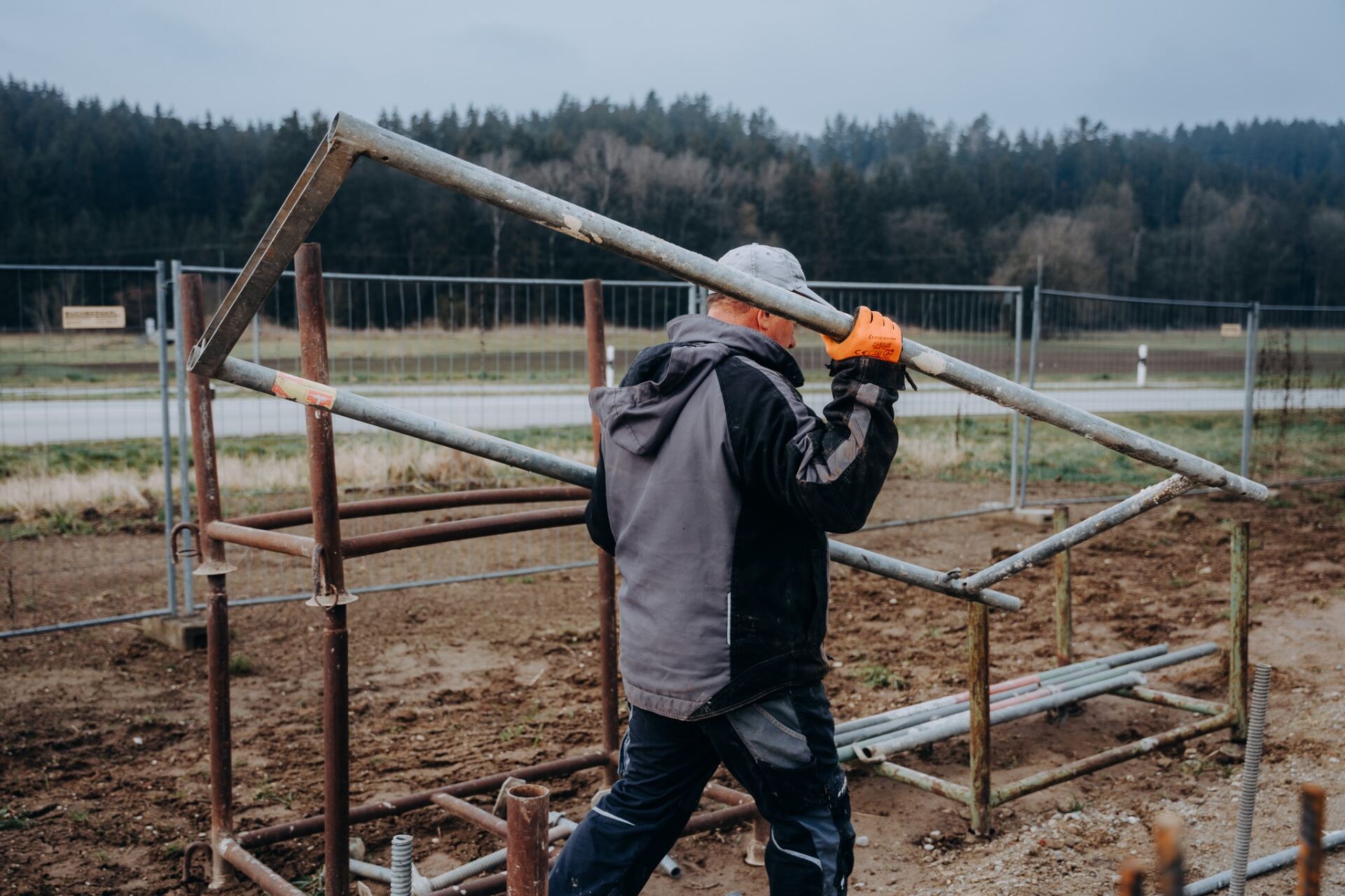 Krojer Bau – Mitarbeiter trägt Gerüstrahmen auf der Schulter über Feld