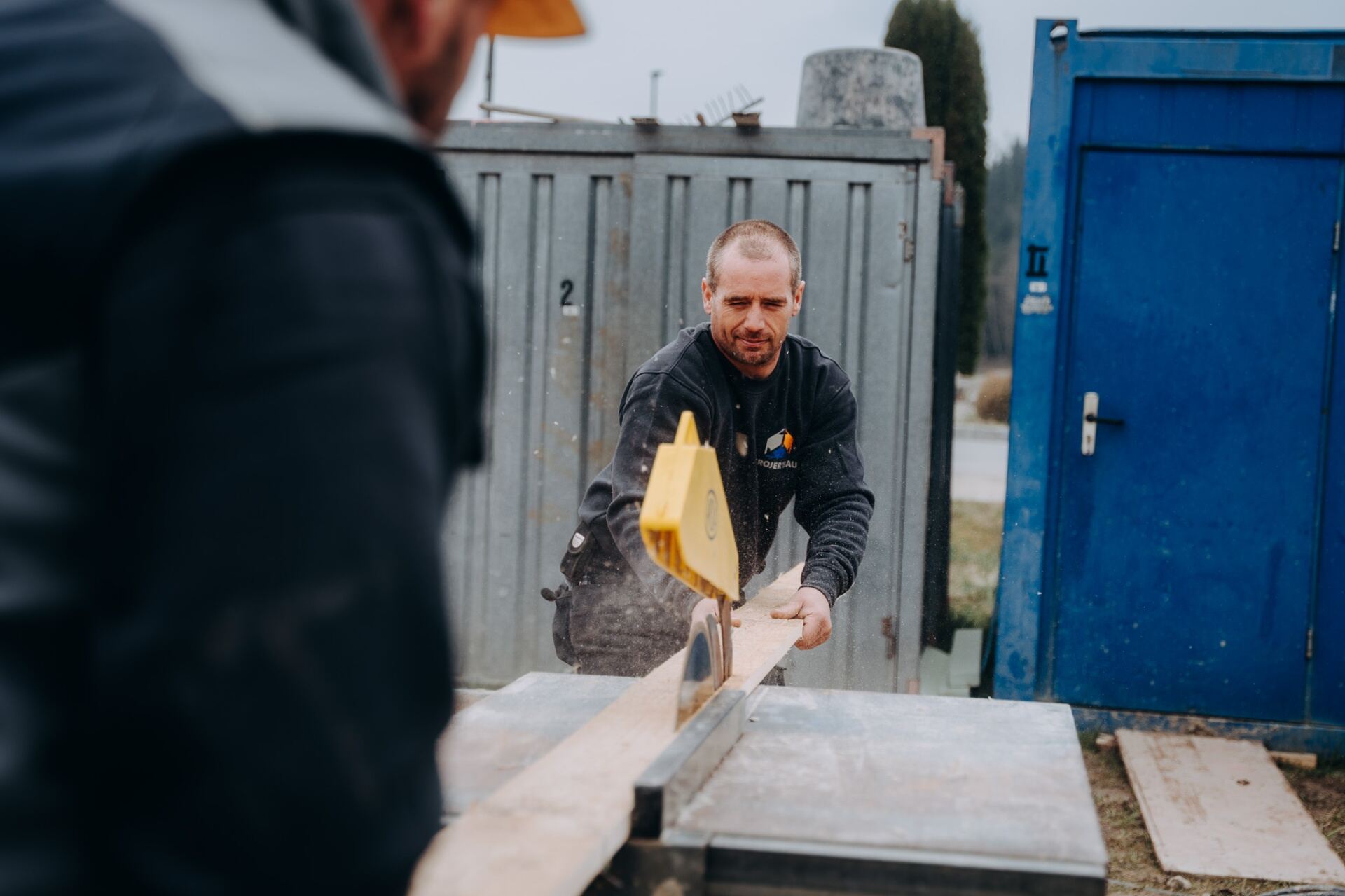 Krojer Bau – Krojer Bau Mitarbeiter an der Tischkreissäge vor Baucontainer